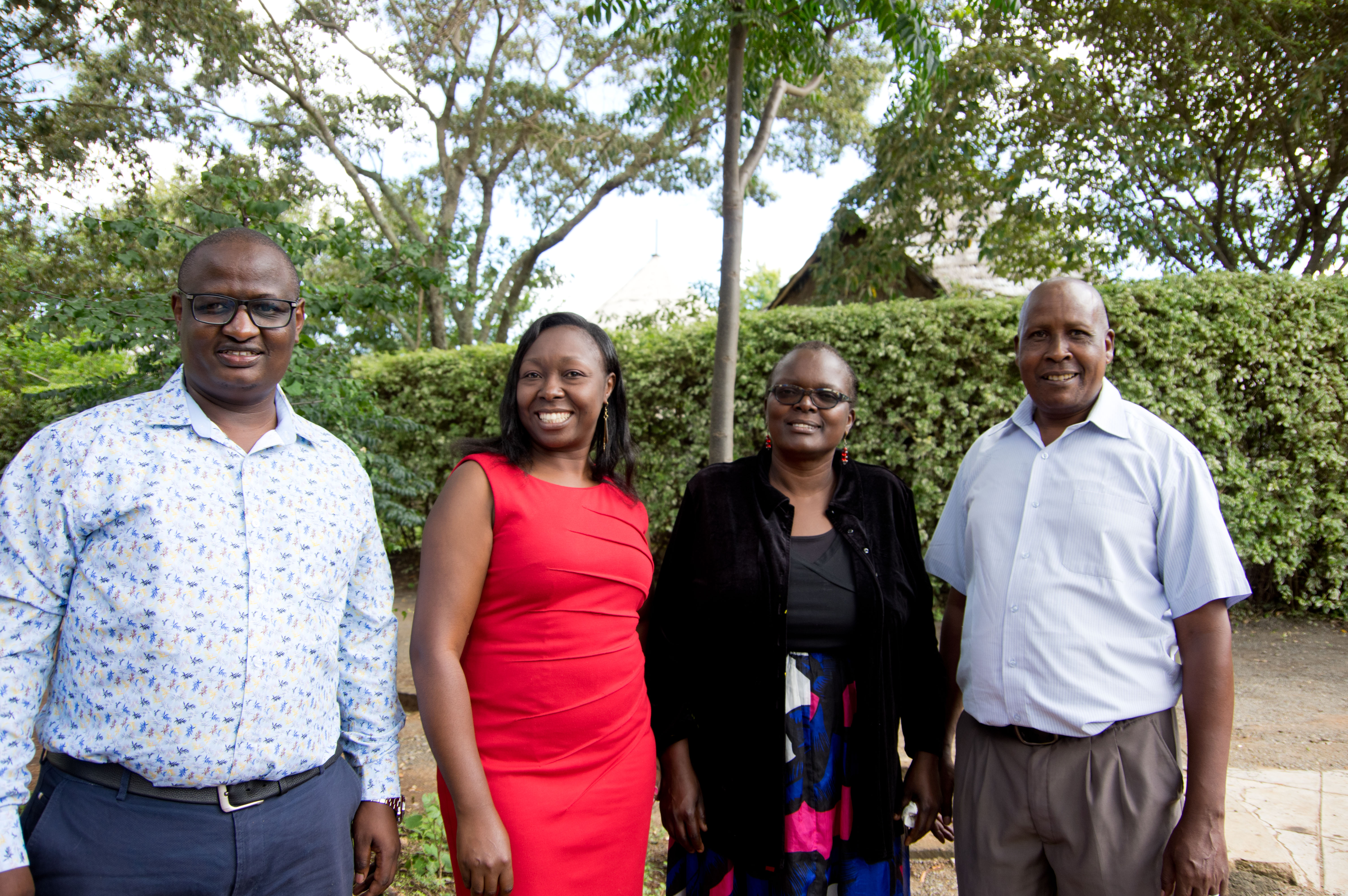 Four Kenyan adults smiling, two men and two women. They are outside and there is a backdrop of trees.